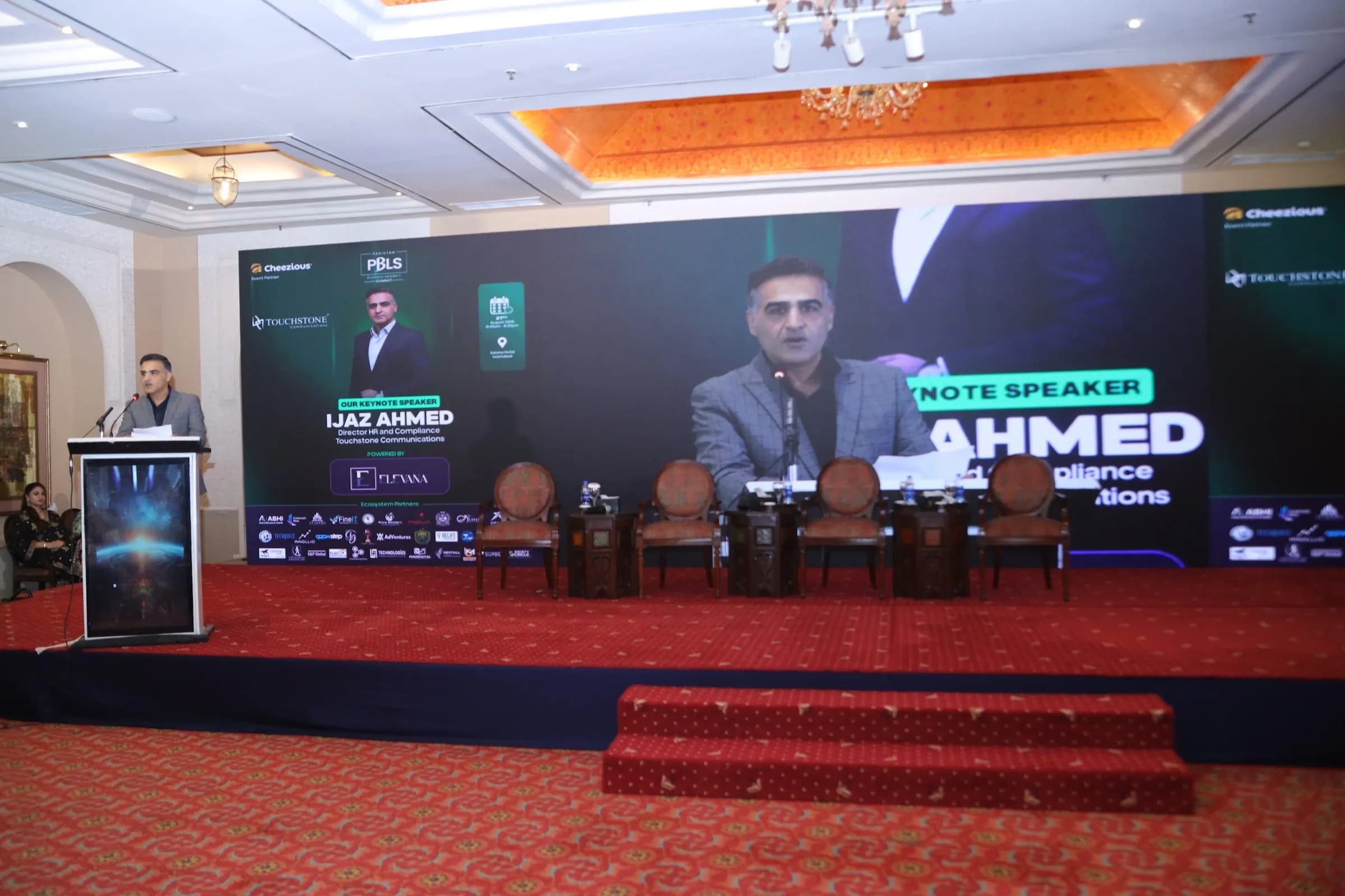 A keynote speaker at the podium, shown on the main LED screen at PBLS Serena Islamabad